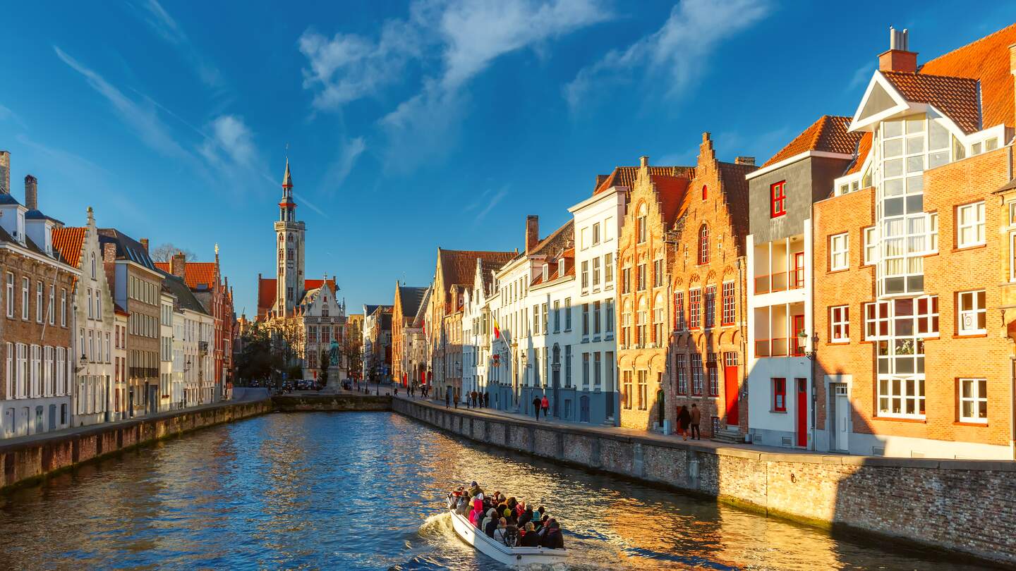 Auf einem Kanal in Bruegge faehrt ein Ausflugsboot. Die historischen Haeuser werden von der Sonne angestrahlt. | © Gettyimages.com/KavalenkavaVolha
