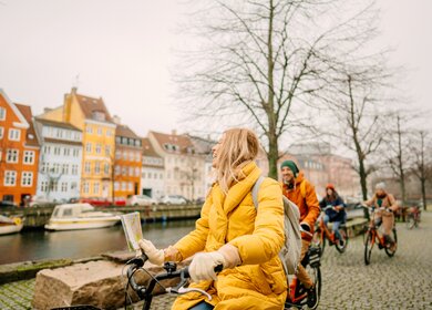 Gruppe Touristen mit dicken Jacken und Mützen auf Fahrrädern am Hafen in Kopenhagen, Dänemark, vor den berühmten Nyhavn | © GettyImages.com/AleksandarNakic