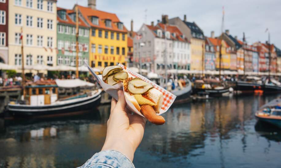 Persoenliche perspektivische Aufnahme der Hand einer Frau, die einen Hot Dog in Kopenhagen, Daenemark, vor den Hafen der Bruecken Strassenkueche haet | © GettyImages.com/Xsandra