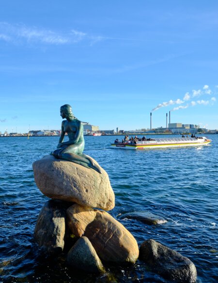 Statue der kleinen Meerjungfrau in Kopenhagen mit Touristenboot und Verbrennungsanlage im Hintergrund | © Gettyimages.com/Angelafoto