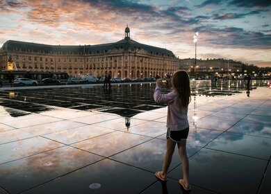 Mädchen beim Fotografieren des berühmten Wasserspiegels von Bordeaux mit dem Place de la Bourse im Hintergrund bei Sonnenuntergang. | © GettyImages.com/	DEBOVE SOPHIE