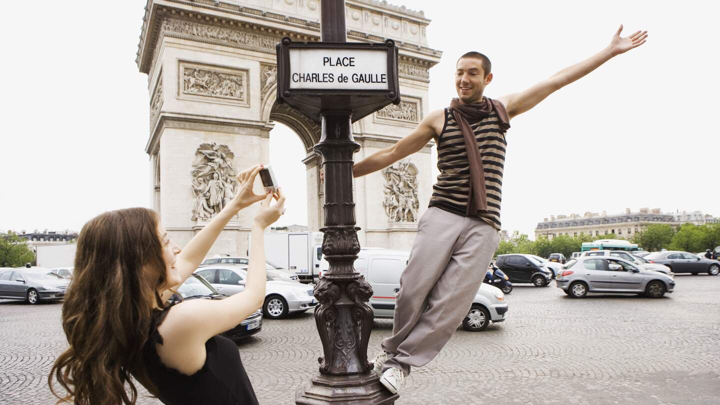 Frau nimmt vor dem Arc de Triomphe ein Foto von einem Mann auf, der auf einem Straßenschild steht.  | © Gettyimages.com/Jupiterimages