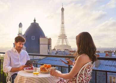 Paar beim Fruehstueck mit Blick auf den Eiffelturm in Paris | © Gettyimages.com/PeopleImages Aufnahme eines jungen Paares beim Fruehstueck auf dem Balkon einer Wohnung mit Blick auf den Eiffelturm in Paris | © Gettyimages.com/PeopleImages