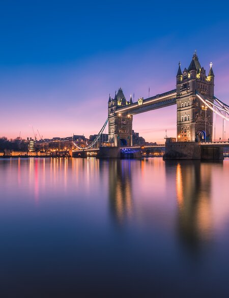Tower Bridge in London bei blauem und rosa Abendhimmel, mit beleuchteten Türmen und ruhigem Wasser, das die Brücke und Lichter reflektiert. | © Gettyimages.com/	Trung Pham