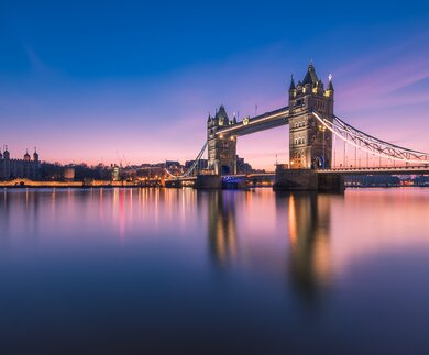 Tower Bridge in London bei blauem und rosa Abendhimmel, mit beleuchteten Türmen und ruhigem Wasser, das die Brücke und Lichter reflektiert. | © Gettyimages.com/	Trung Pham