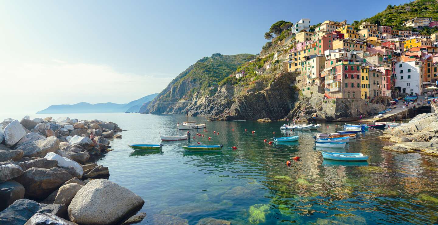 Idyllischer Blick vom smaragdgruenen Mittelmeer auf die Stadt Riomaggiore im Cinque Terre Nationalpark.  Die bunten Häuser des Dorfes und der Hafen strahlen im Sonnenlicht, während kleine Fischerboote auf dem Wasser vor der Küste treiben. | © Gettyimages.com/spooh