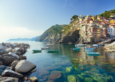 Idyllischer Blick vom smaragdgruenen Mittelmeer auf die Stadt Riomaggiore im Cinque Terre Nationalpark.  Die bunten Häuser des Dorfes und der Hafen strahlen im Sonnenlicht, während kleine Fischerboote auf dem Wasser vor der Küste treiben. | © Gettyimages.com/spooh