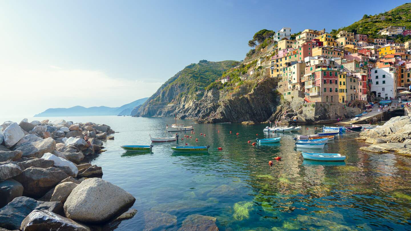 Idyllischer Blick vom smaragdgruenen Mittelmeer auf die Stadt Riomaggiore im Cinque Terre Nationalpark.  Die bunten Häuser des Dorfes und der Hafen strahlen im Sonnenlicht, während kleine Fischerboote auf dem Wasser vor der Küste treiben. | © Gettyimages.com/spooh