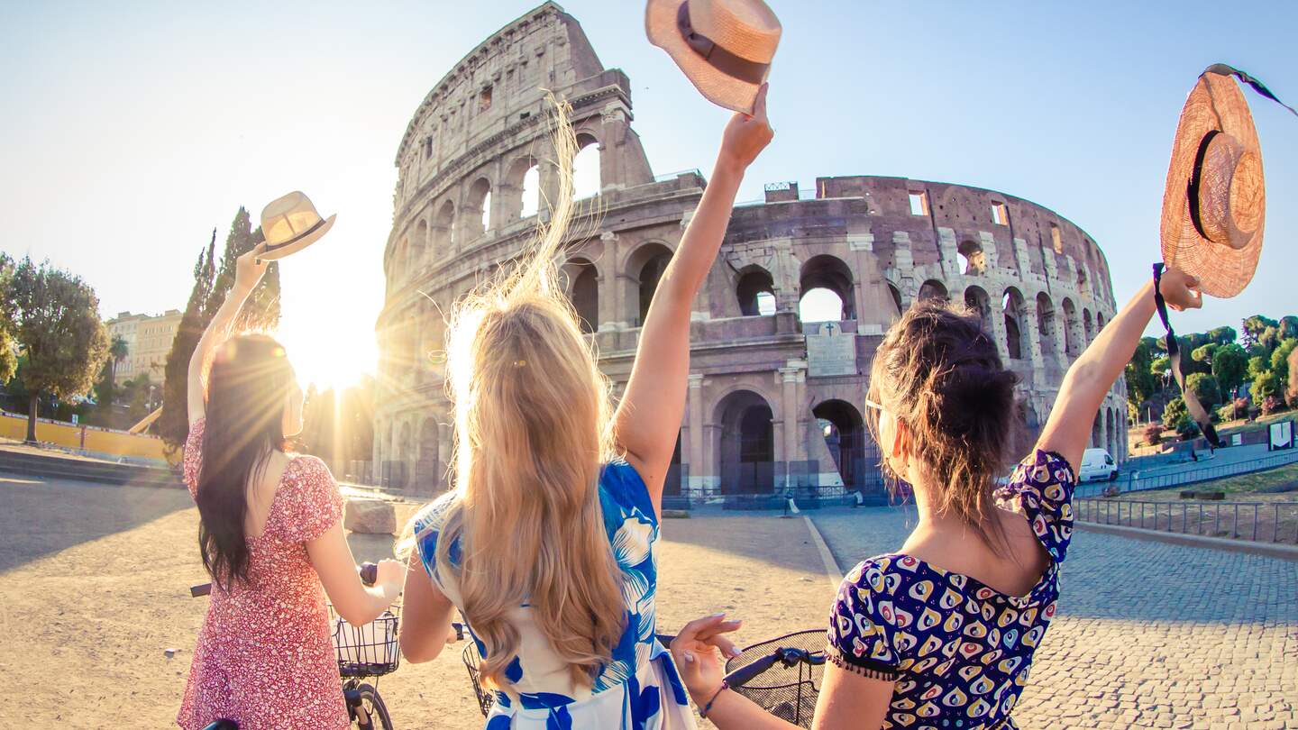 Drei glueckliche junge Frauen freundet Touristen mit Fahrrädern winken Huete am Kolosseum in Rom, Italien bei Sonnenaufgang | © Gettyimages.com/tfilm