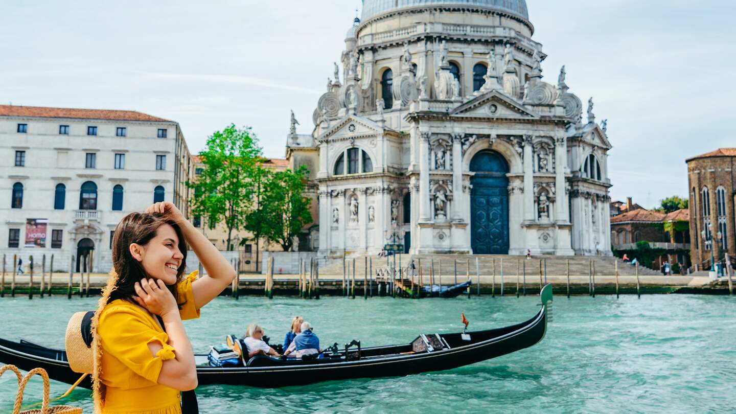 Frau im gelben Sonnenkleid sitzt am Ufer mit Blick auf den grossen Kanal in Venedig. | © Gettyimages.com/Vera_Petrunina