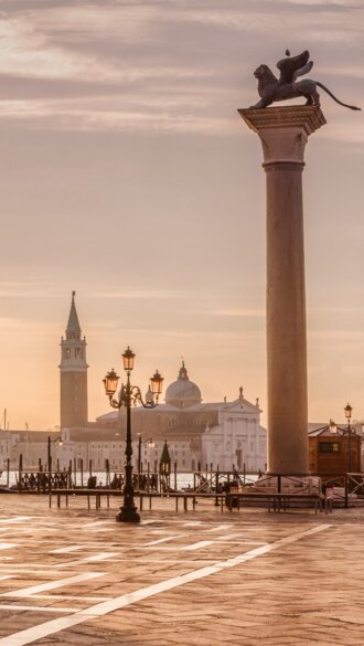 Auf dem Markusplatz in Venedig laufen Menschen. Die Sonne scheint durch den Dogenpalast. | © Gettyimages.com/tunart