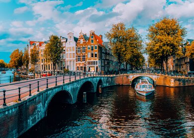 Ansicht von Amsterdam mit einem Kanal, auf dem ein Boot faehrt, gewoelbten Bruecken und traditioneller niederlaendischer Architektur unter einem blauen Himmel mit vereinzelten Wolken.  | ©  Gettyimages.com/miniloc