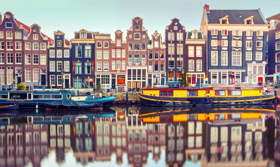 Amsterdams traditionelle Haeuserarchitektur mit Hausbooten spiegelt sich am fruehen Morgen in einer Gracht | © Gettyimages.com/KavalenkavaVolha