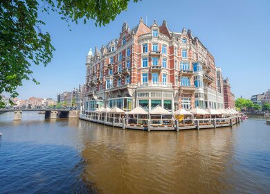 Gracht in Amsterdam mit verschnoerkelten Gebaeuden. Vor dem Gebaeude befindet sich ein Café mit Sonnenschirmen. | ©  Gettyimages.com/David Pedre