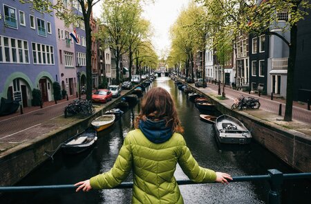 Junge Frau in hellgruener, taillerter Steppjacke steht auf einer Grachtenbrücke in Amsterdam und blickt auf den Kanal | © Gettyimages.com/bortnikau
