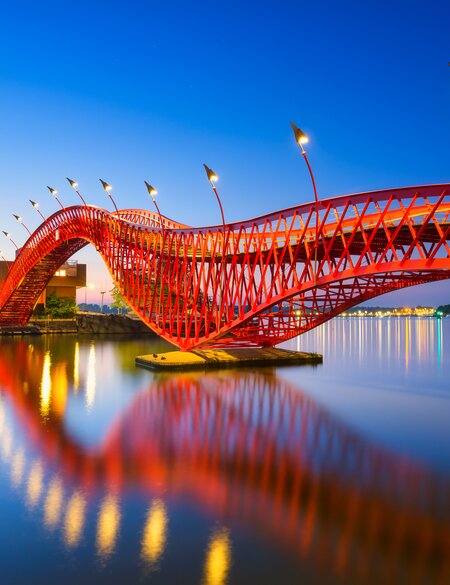 Die abendlich kunstvoll beleuchtete Fußgaengerbrücke Python Bridge, auch High Bridge genannt, in Amsterdam spiegelt sich im Kanal | © Gettyimages.com/Biletskiy_Evgeniy
