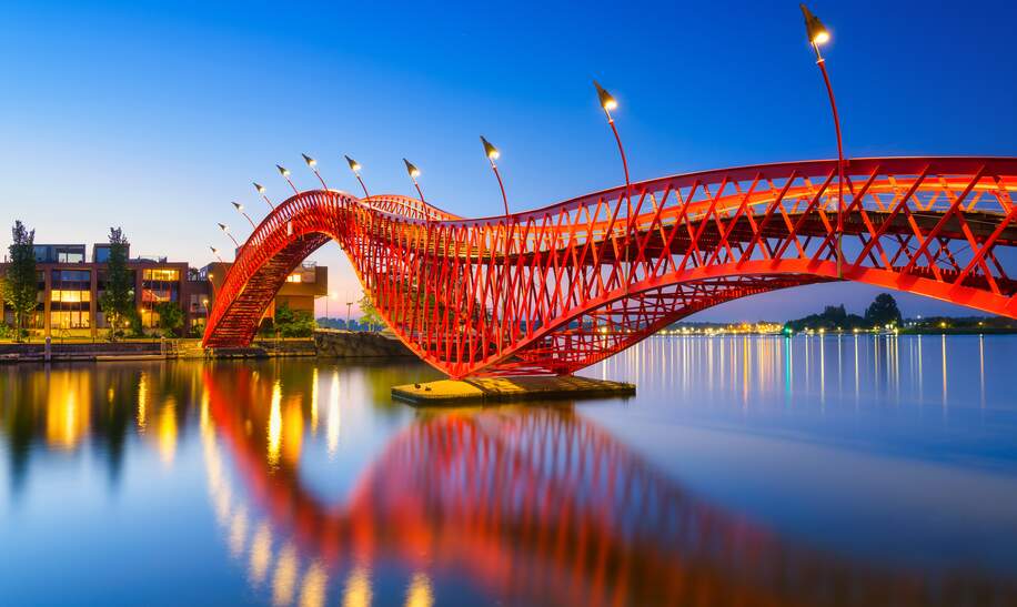Die abendlich kunstvoll beleuchtete Fußgaengerbrücke Python Bridge, auch High Bridge genannt, in Amsterdam spiegelt sich im Kanal | © Gettyimages.com/Biletskiy_Evgeniy