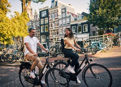 Paar faehrt auf Fahrraedern vorbei an Grachten der Strassen von Amsterdam | © GettyImages.com/pixdeluxe