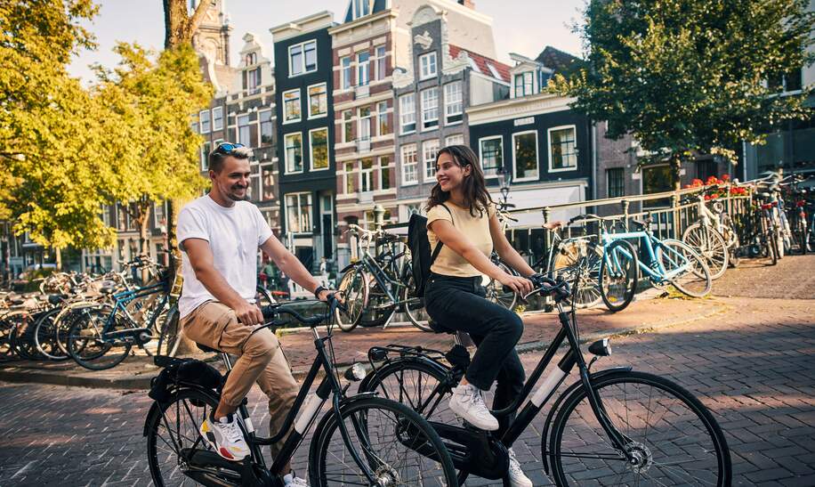 Paar faehrt auf Fahrraedern vorbei an Grachten der Strassen von Amsterdam | © GettyImages.com/pixdeluxe