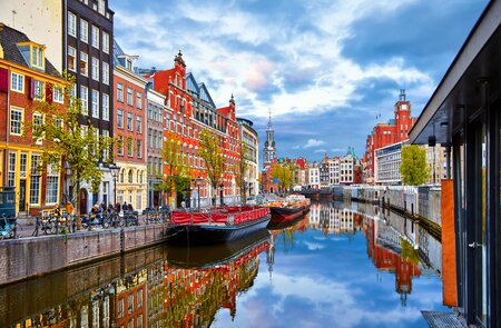 Bunte Häuser und Boote entlang eines Kanals in Amsterdam, mit Spiegelungen im ruhigen Wasser und blauem Himmel mit Wolken im Hintergrund. | © Gettyimages.com/	Yasonya