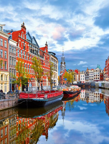 Bunte Häuser und Boote entlang eines Kanals in Amsterdam, mit Spiegelungen im ruhigen Wasser und blauem Himmel mit Wolken im Hintergrund. | © Gettyimages.com/	Yasonya