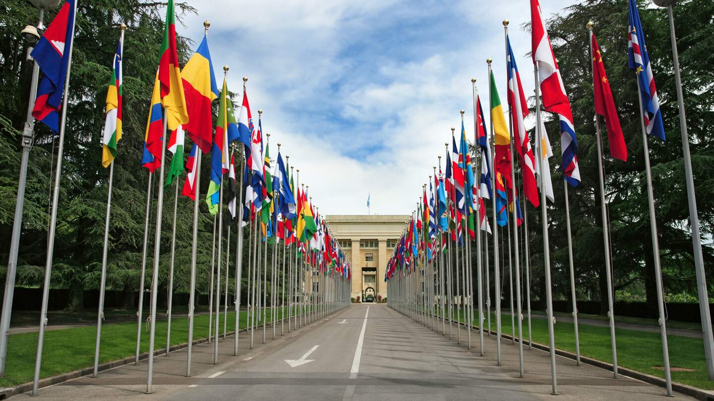 Eingang und Gebaeude der Vereinten Nationen in Genf mit Flaggen entlang des Weges | © Gettyimages.com/sumnersgraphicsinc
