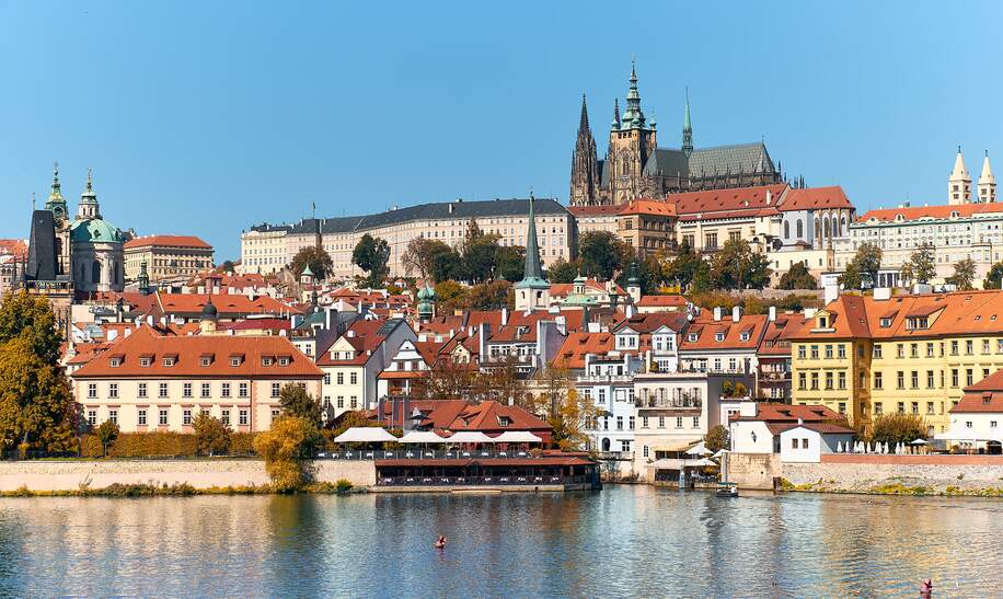 Prager Burg mit orangefarbenen Dächern historischer Gebäude von Mala Strana, die sich an einem hellen Sommertag in Prag im Flusswasser spiegeln. | © Gettyimages.com/anyaivanova