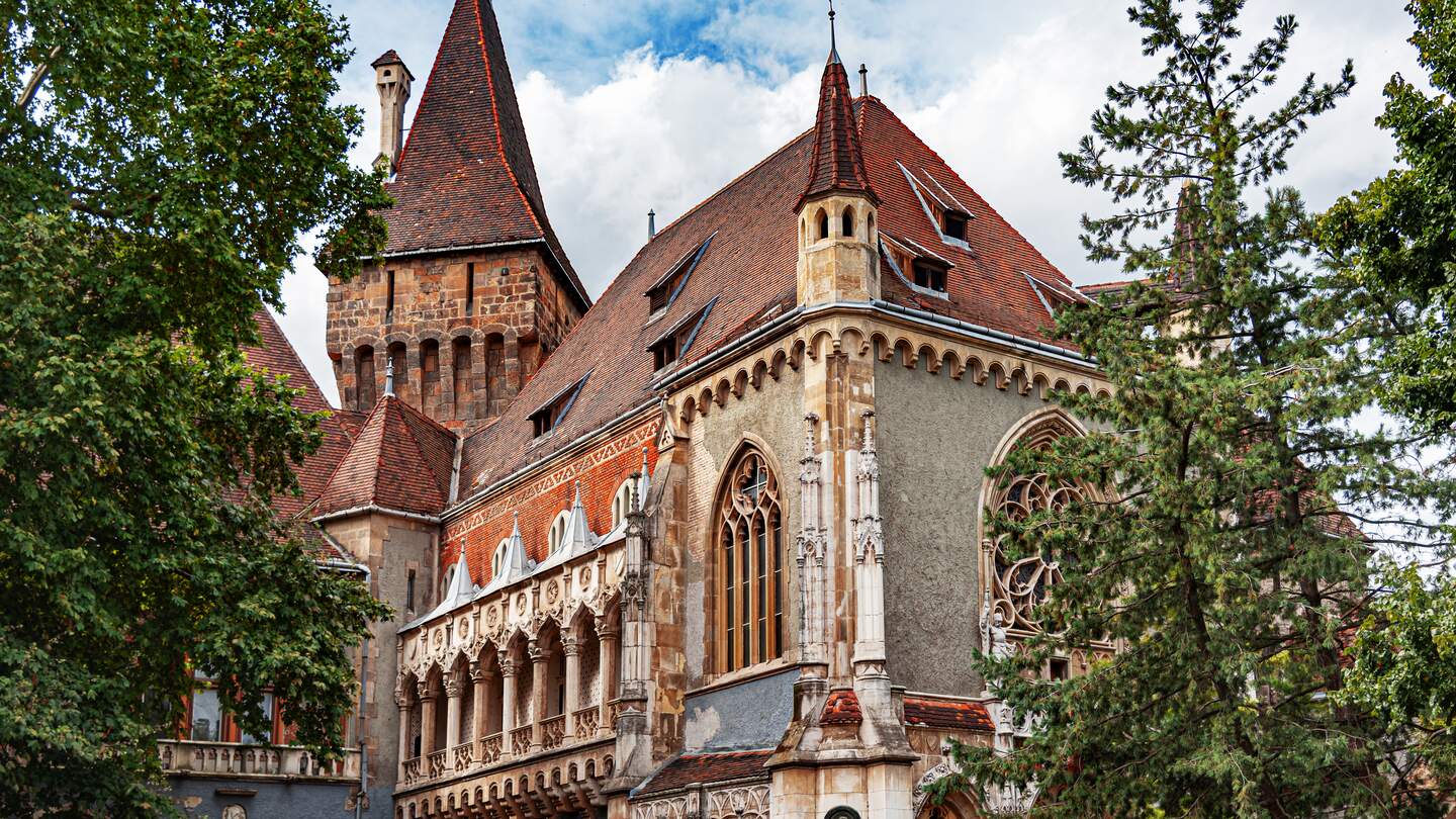 Vajdahunyad-Schloss in Budapest, Ungarn, mit roten Ziegeldächern, Türmen und reich verzierten gotischen Fenstern, umgeben von grünen Bäumen unter einem teilweise bewölkten Himmel. | © Gettyimages.com/Believe_In_Me