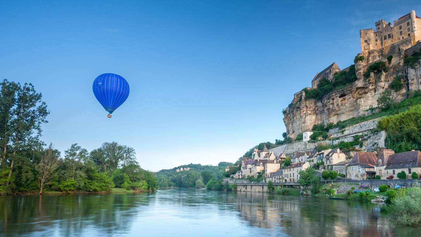 Chateaux Beynac auf einer Klippe, auf der linken Seite fliegt ein blauer Heissluftballon | © Gettyimages.com/garethkirklandphotogrphy