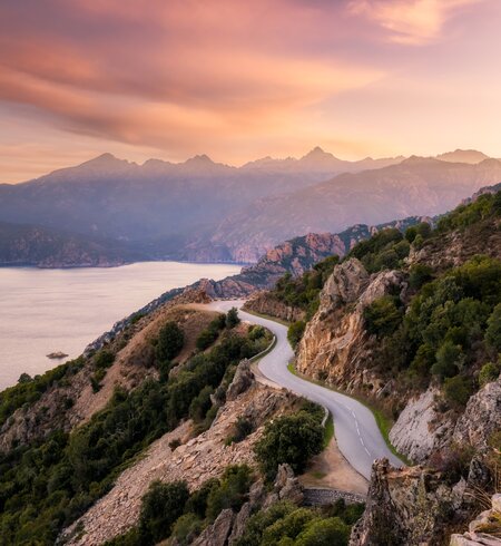Die Straße D824 schlängelt sich von Capu Rossu entlang der Küste in Richtung Piana an der Westküste Korsikas, während die frühe Morgensonne die fernen Berge beleuchtet | © Gettyimages.com/77joningall