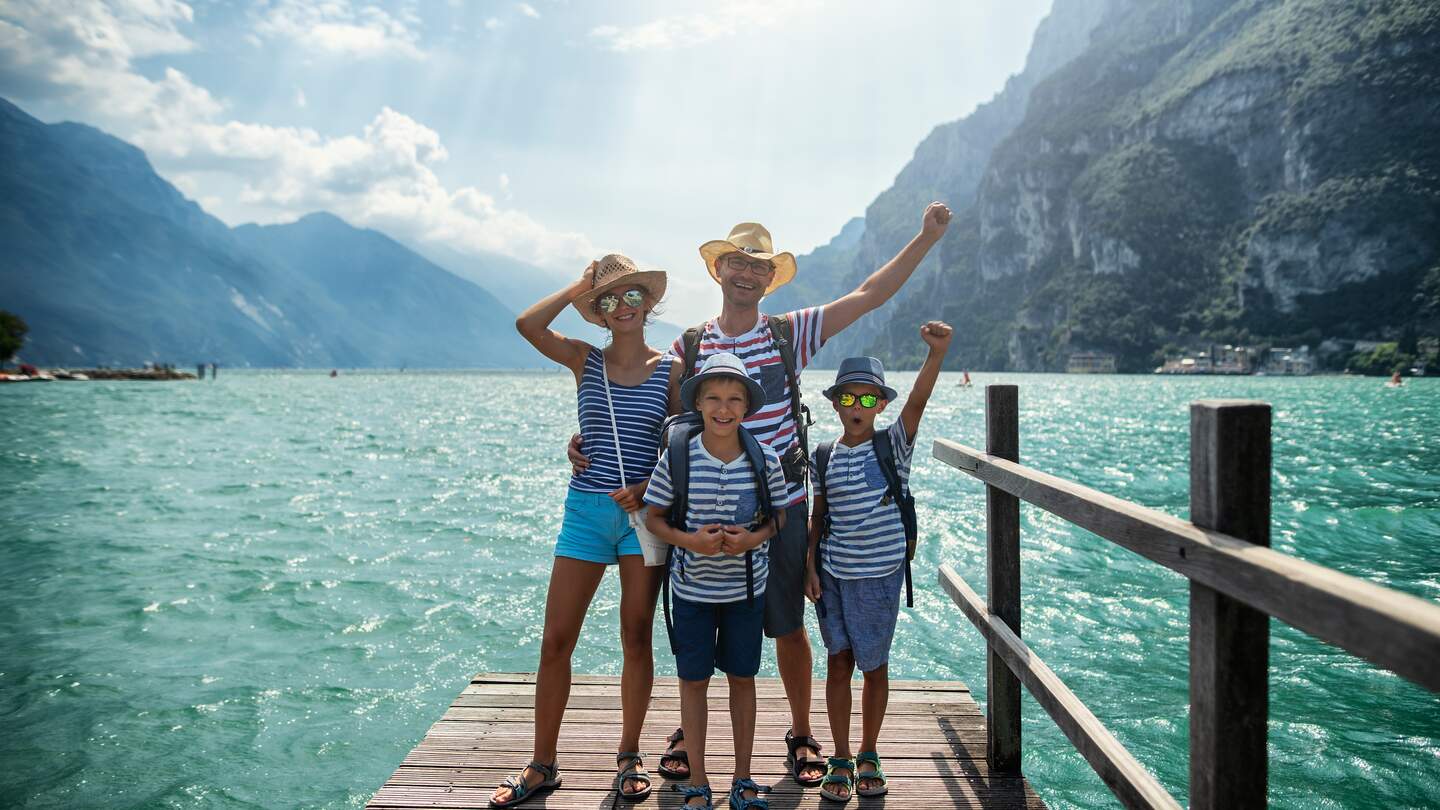 Familie geniesst Urlaub in Italien. Vater und Kinder stehen auf einem Pier in Riva del Garda und jubeln in die Kamera. Dahinter bietet sich ein herrlicher Blick auf den Gardasee, umgeben von den Alpen | © Gettyimages.com/Imgorthand