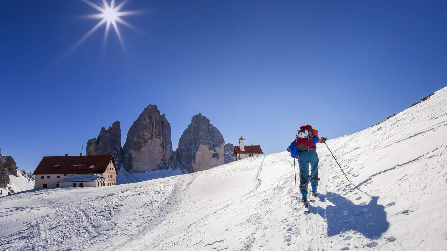 Skifahrer laeuft einen schneebedeckten Berg hoch bei strahlendem Sonnenschein, der Gebirgsstock Drei Zinnen ist im Hintergrund zu sehen | © Gettyimages.com/DieterMeyrl