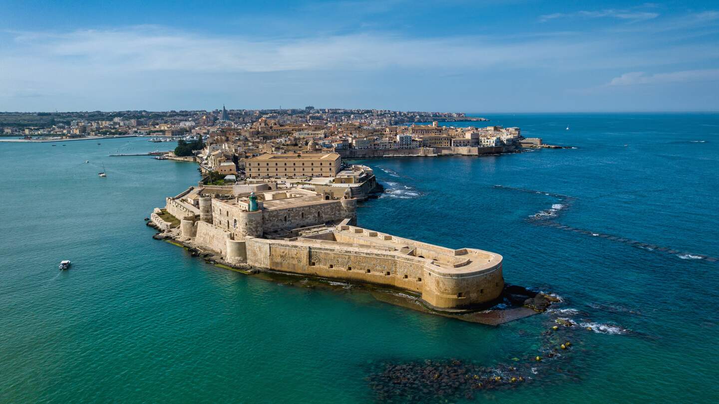 Drohnenblick auf die "krokodilkopffoermige" Insel Ortygia, die mit der Stadt Syrakus verbunden ist, mit Maniace Castello an der Ostkueste der Insel Sizilien in Richtung Mittelmeer unter blauem Sommerhimmel. Syrakus. Festung Maniace, Insel Ortygia, Sizilien, Italien, Suedeuropa | © Gettyimages.com/mlenny