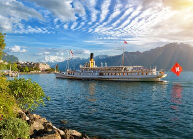 Wunderschoene Sommerabendlandschaft des Genfersees mit malerischen Ufern und Ausflugsschiff gegen alpine Berge in den Strahlen der untergehenden Sonne in Montreux, Schweiz | © Gettyimages.com/connect11