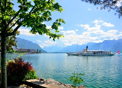 Blick von Montreux auf den Genfersee mit vorbeifahrendem Ausflugsschiff bei Sonnenschein und leichten Wolken | © Gettyimages.com/alxpin