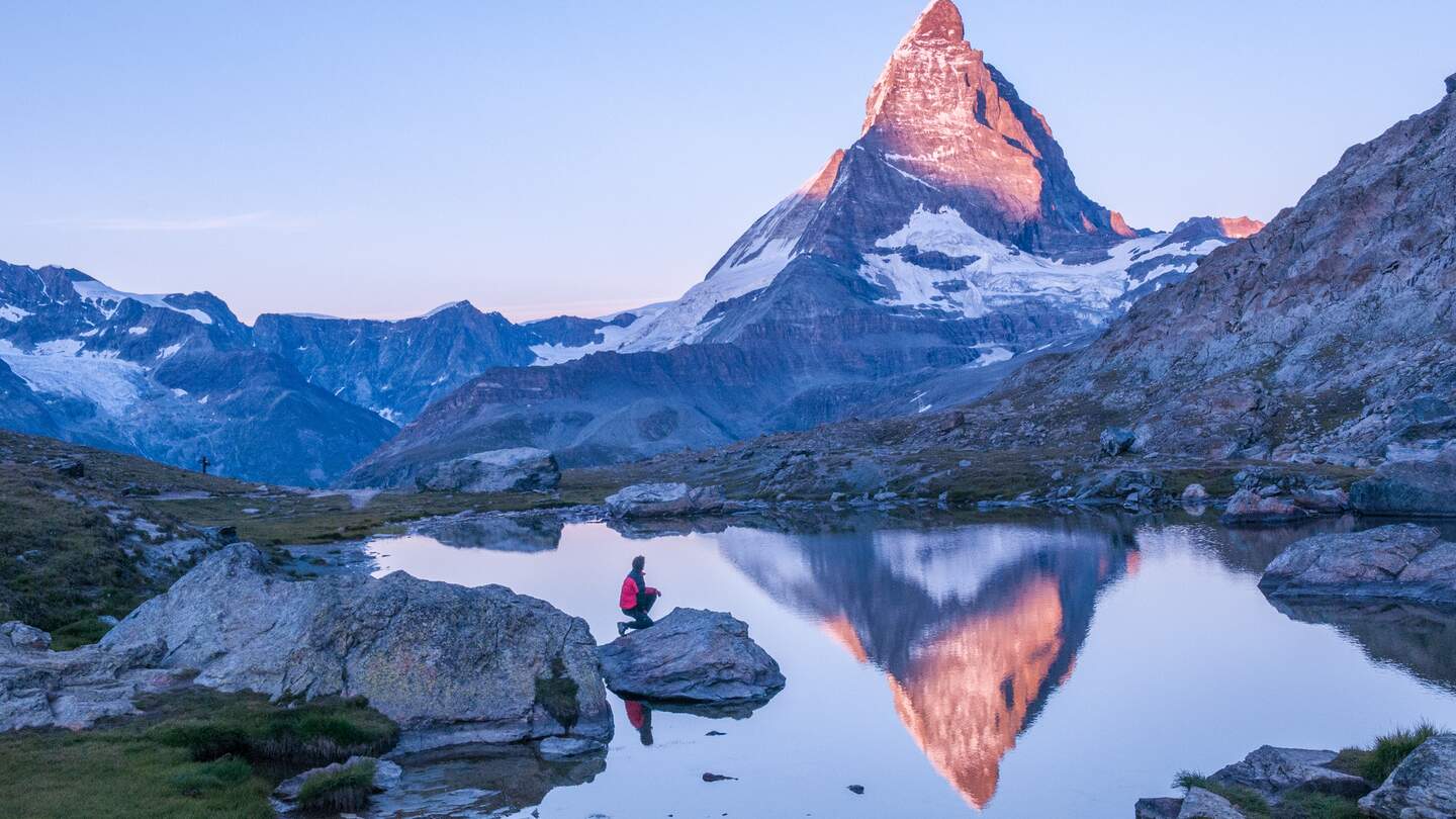 Fruehmorgens Sonnenaufgangsszene auf dem Matterhorn, das sich rosa im See spiegelt, mit Mann auf einem Felsen mit roter Daunenjacke und klarem blauen Himmel  | © Gettyimages.com/wilpunt