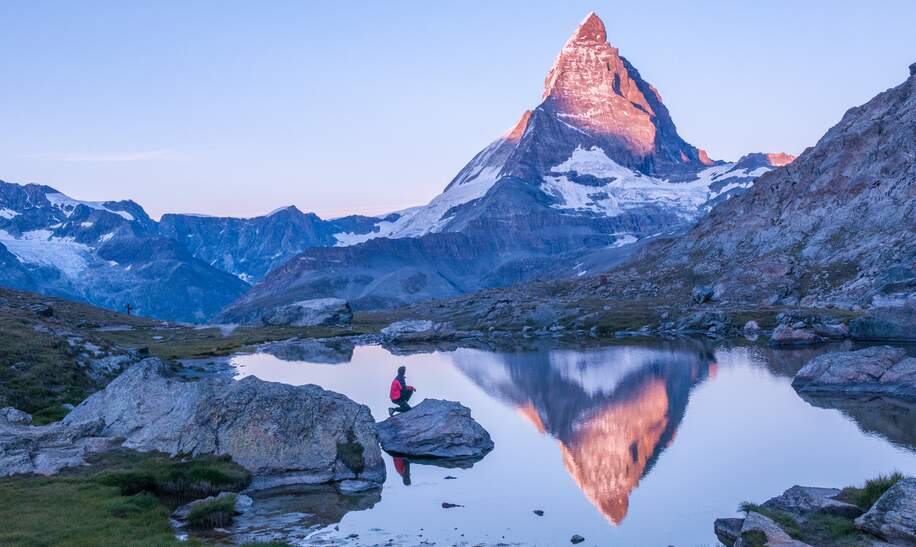 Frühmorgens Sonnenaufgangsszene auf dem Matterhorn, das sich rosa im See spiegelt, mit Mann auf einem Felsen mit roter Daunenjacke und klarem blauen Himmel  | © Gettyimages.com/wilpunt