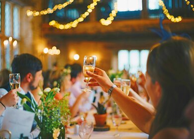Mehrere Personen stoßen mit Sektgläsern an einer festlich dekorierten Tafel in einem warm beleuchteten Saal mit Lichterketten an. | © Foto von Al Elmes auf Unsplash