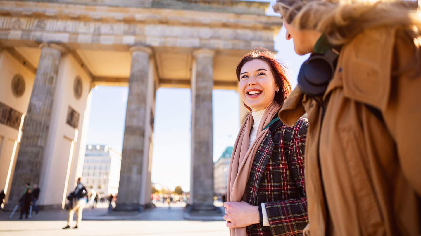 Zwei Personen stehen vor dem Brandenburger Tor in Berlin und unterhalten sich bei sonnigem Wetter im Freien. | © GettyImages.com/	SrdjanPav