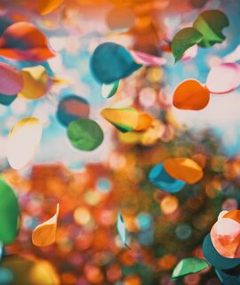 Mehrfarbige Konfettikreise schweben durch die Luft vor einem farbenfrohen, weich verschwommenen Hintergrund mit Bokeh-Effekt. | © GettyImages.com/melis82