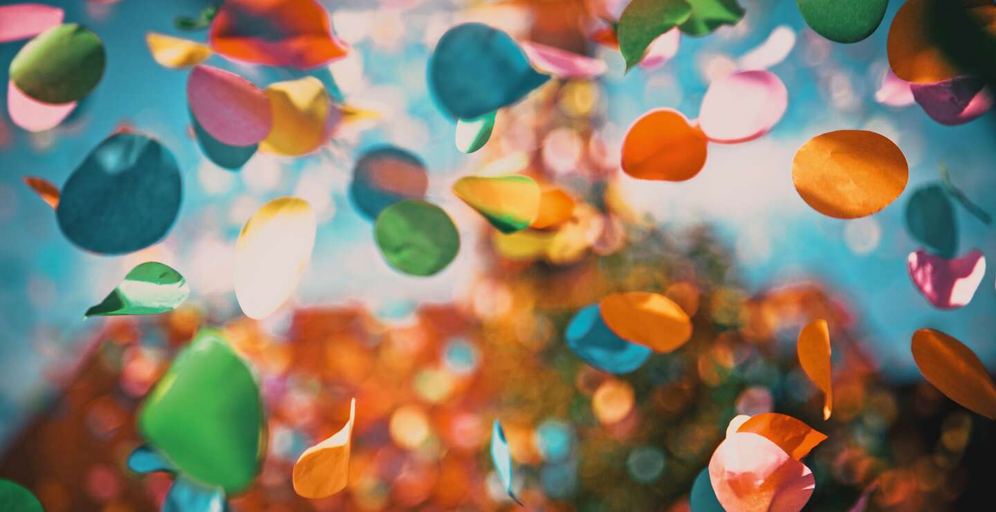 Mehrfarbige Konfettikreise schweben durch die Luft vor einem farbenfrohen, weich verschwommenen Hintergrund mit Bokeh-Effekt. | © GettyImages.com/melis82