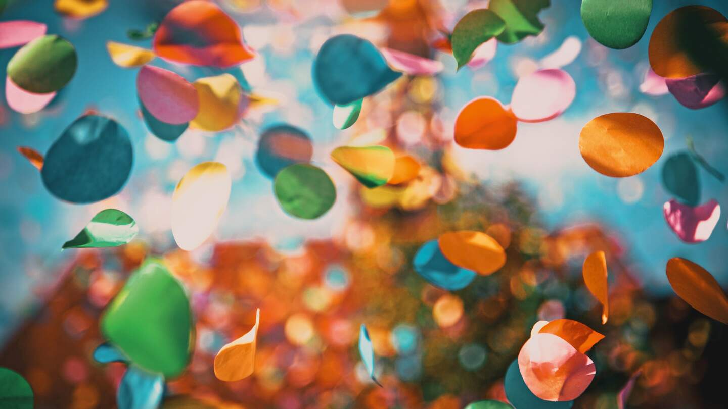 Mehrfarbige Konfettikreise schweben durch die Luft vor einem farbenfrohen, weich verschwommenen Hintergrund mit Bokeh-Effekt. | © GettyImages.com/melis82