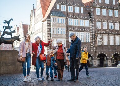 Das Bild zeigt eine lebendige Szene auf dem Marktplatz der Stadt Bremen, die von historischer Architektur und alltäglichem Leben geprägt ist. Im Vordergrund bewegt sich eine Gruppe von Personen über das charakteristische Kopfsteinpflaster, während im Hintergrund eindrucksvolle Gebäude im Stil der Weser-Renaissance mit ihren aufwendig verzierten Fassaden und zahlreichen Fenstern zu sehen sind. Auf der linken Seite des Bildes steht die bekannte Skulptur der Bremer Stadtmusikanten, die mit den übereinandergestellten Tieren Esel, Hund, Katze und Hahn eines der bekanntesten Wahrzeichen der Stadt darstellt. Die gesamte Szene vermittelt den Eindruck eines entspannten Stadtbummels in einer geschichtsträchtigen Umgebung, in der sich kulturelles Erbe und modernes Leben auf natürliche Weise miteinander verbinden. | © WFB / Katja Thiele