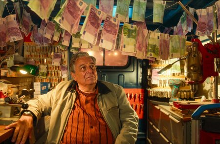 Der Schauspieler Christian Clavier steht in dem Kinofilm "Ab durch die Mitte – Mit Vollgas durch Paris" in einem Werkzeugraum mit neutralem Gesichtsausdruck und über ihm hängen viele Geldscheine | © Happy Entertainment