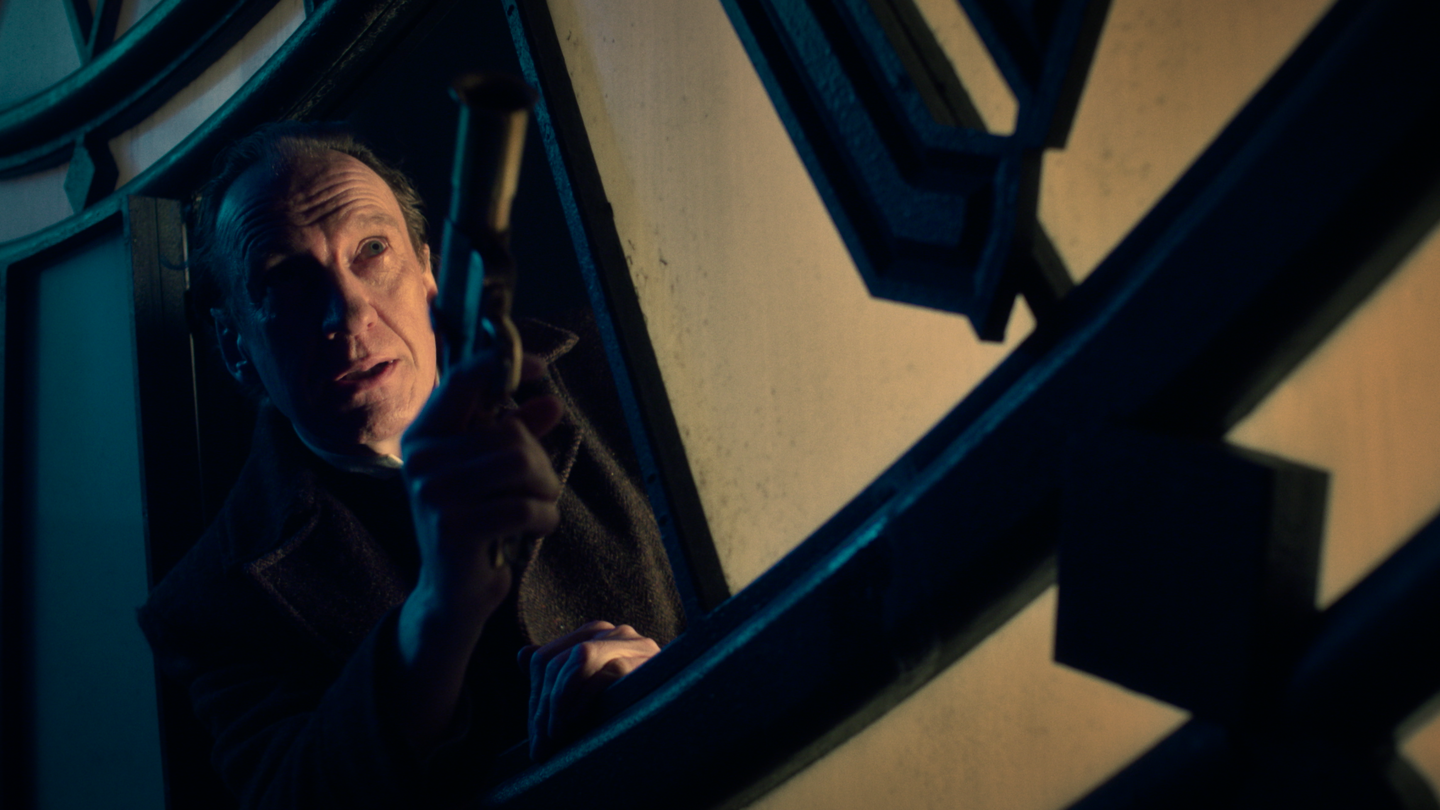 Eine Person steht in einem dunklen Raum vor einem großen, geometrisch gestalteten Metall- oder Glasstruktur-Element, das an ein Uhrwerk erinnert.  | © polyband/Palatin Media/Albion Television Ltd