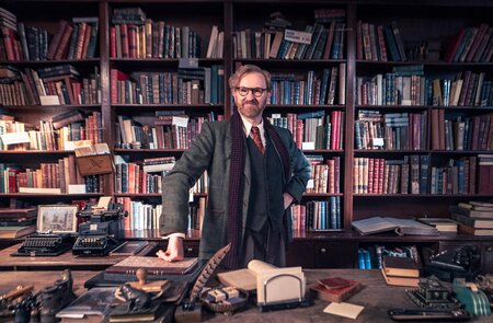 Die Figur Gabriel Book aus der Serie "Bookish" steht lächelnd vor seiner Buchsammlung in seinem Antiquariat | © polyband/Beta Film/UK TV