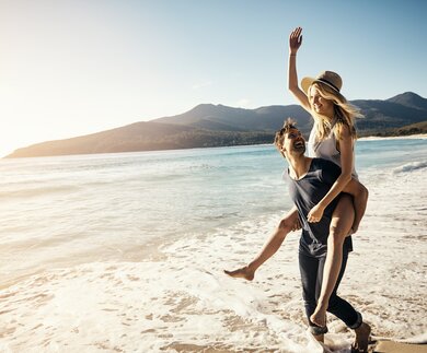 Ein junger Mann trägt eine junge Frau Huckepack an einem Strand im Sommer | © HanseMerkur Reiseversicherung/pixdeLuxe