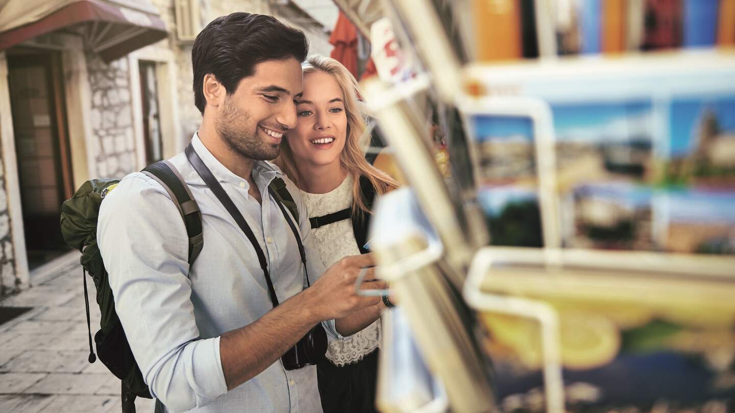 Ein junges Paar schaut sich Postkarten an einem Postkartenständer in einer Stadt an | © HanseMerkur Reiseversicherung