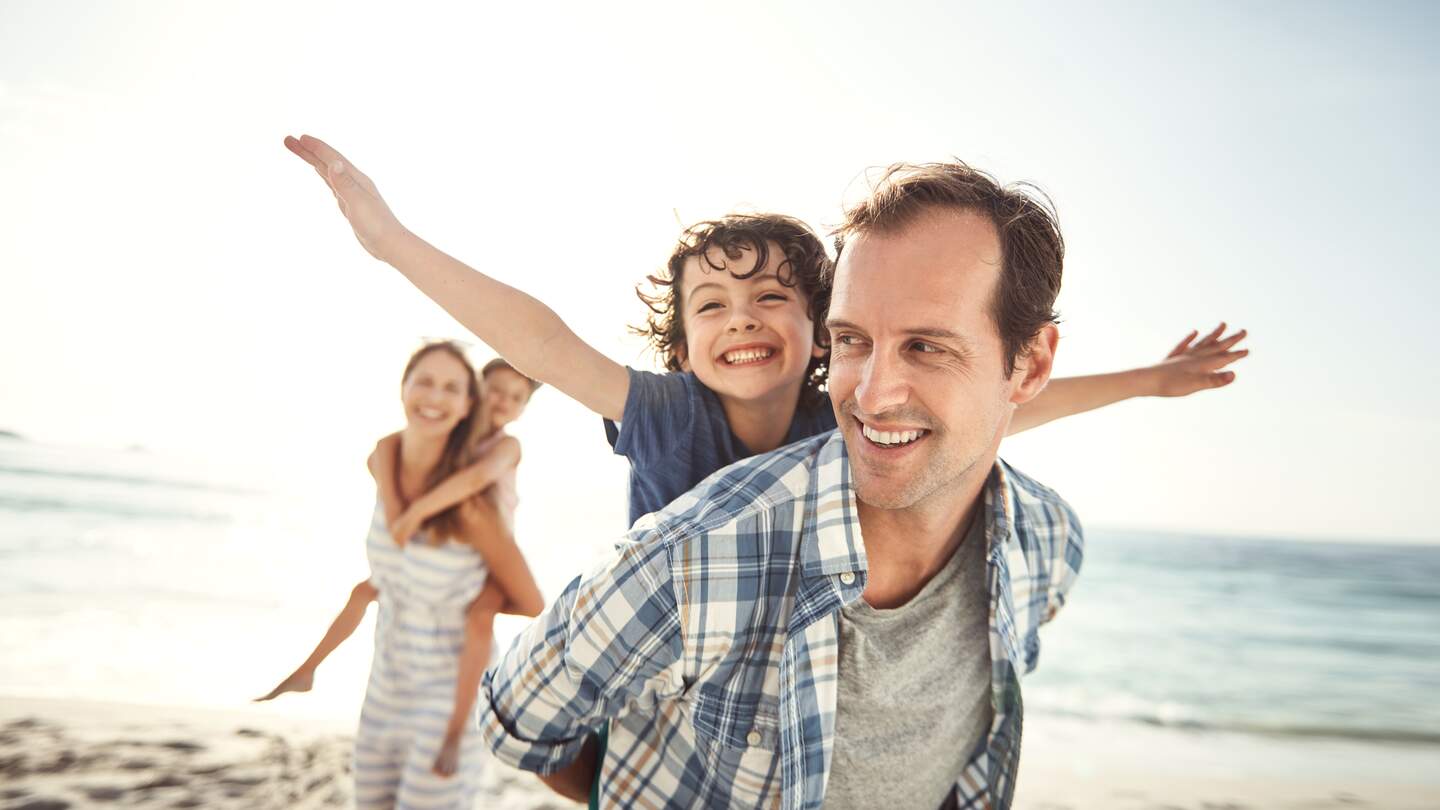 Eine Familie mit zwei Kindern laufen ausgelassen an einem Stand im Sommer | © HanseMerkur Reiseversicherung
