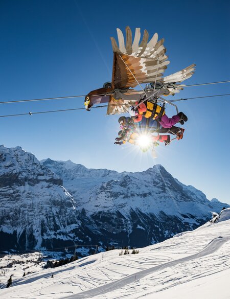 First Glider | © Jungfraubahnen, David Birri Gruppe von Personen, die an einem gleiterförmigen Fahrgeschäft in Form eines großen Vogels über eine verschneite Berglandschaft schweben. Der Gleiter ist an Seilen befestigt, die auf ein Abenteuer- oder Freizeitpark-Erlebnis hindeuten. Die Sonne scheint hell hinter der Gruppe und taucht die schneebedeckten Berge in dramatisches Licht | © Jungfraubahnen, David Birri
